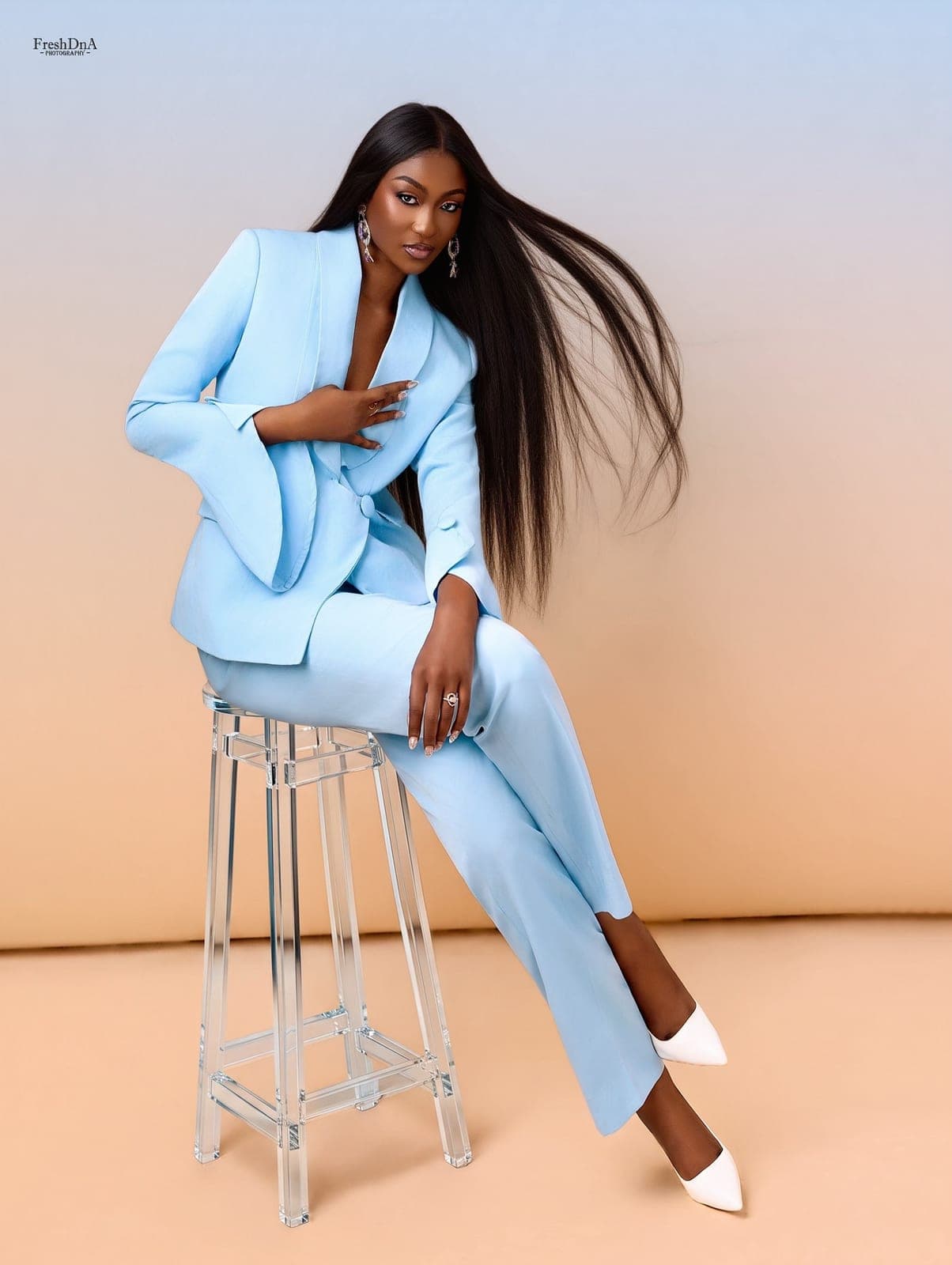 Woman in a tailored sky-blue Moirai power suit seated on a clear stool against a peach backdrop